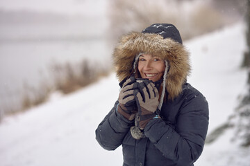 Happy smiling woman wearing a fur trimmed hooded jacket enjoying a mug of coffee cradled in her gloved hands outdoors in winter snow in a concept of the seasons in a close up portrait