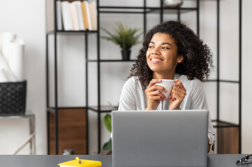 Smiling young joyful African American female freelancer enjoying morning coffee while working on...
