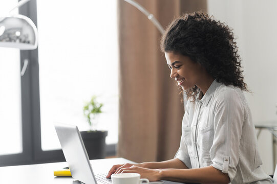 Side view portrait of a positive inspired African American female entrepreneur businesswoman sitting at the desk and typing on a laptop, writing article, working on an online virtual project from home