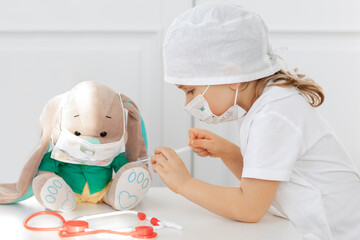 Little child girl makes injection to toy. Cute child with medical mask playing doctor, holding syringe with vaccine. Coronavirus covid vaccination concept. Kid play role game at home.