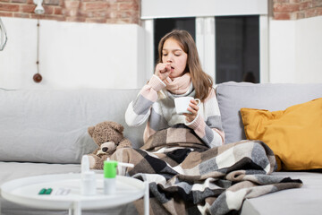 Tonsillitis, influenza, pandemy, illness and healthcare concept. Attractive tired ill teen girl with scarf around neck, sitting on soft couch at home and coughing, drinking hot tea.