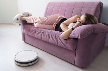 girl sleeps on the couch next to a working white robot vacuum cleaner