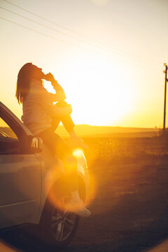 Woman Stop At Roadside To Enjoy Sunset. Sitting On Car Hood