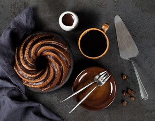 Cake with chocolate icing on a wooden board with a linen napkin, coffee, sauce, plates and a serving spatula on a dark gray background top view