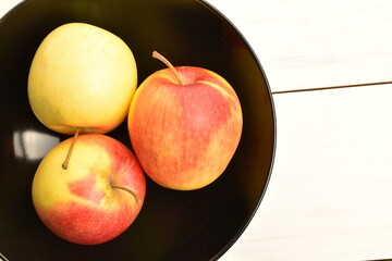 Three ripe organic, juicy, fragrant apples in a black ceramic plate, close-up, on a painted wooden table.