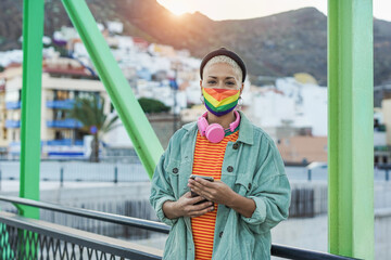 Obraz premium Beautiful short hair woman holing smartphone while wearing lgbt rainbow face mask for coronavirus outbreak