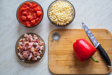 Home made food concept: top view on cutting board with red pepper and a knife, cheese, bacon and tomatoes in the bowls