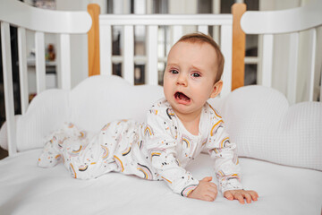 Upset baby in pajama crying. Sad baby in  sleepwear crying loudly while lying in cradle in baby's...