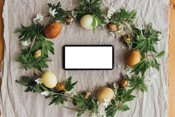 Phone with blank screen in easter rustic wreath with eggs and flowers flat lay. Seasons greetings