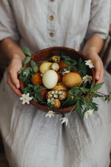 Woman in linen dress holding easter eggs and spring flowers in wooden bowl. Aesthetic holiday