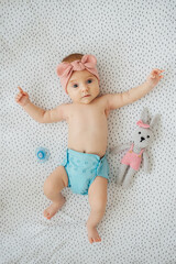 Top view of adorable infant in reusable diaper and stylish headband lying near toy bunny on bed at...