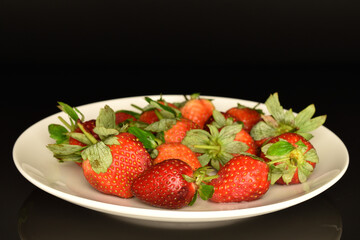 Several berries of bright red ripe organic tasty strawberries on a white ceramic saucer. The background is black.