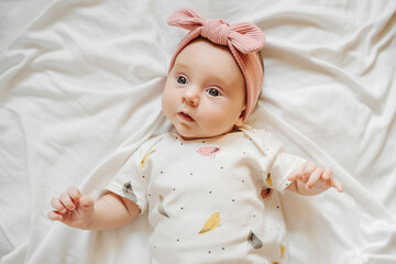 Cute stylish baby in trendy clothes and pale pink headband lying on bed. Fashion baby girl