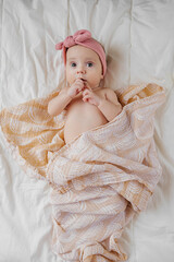 Astonished infant in trendy headband lying on soft mattress wrapped in ornamental coverlet at home. Cute Baby  wrapped in  muslin swaddle blanket on bed - obrazy, fototapety, plakaty