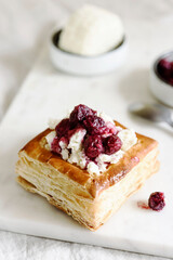 Puff pastry squares with ricotta and cherry sauce on white marble tray. Baked small square cakes topped with cheese and berry sauce