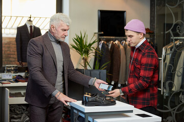 A mature man with gray hair and a sporty physique is applying a cellphone to a point of sale terminal in a clothing store. A male customer with a beard is paying to a shop assistant in a boutique.