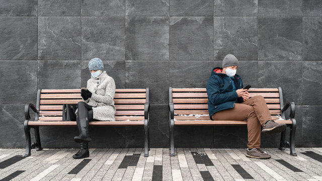 Young caucasian man and woman keep social distance looking at their smartphones and wearing protective face masks. They sit on benches against backdrop of stone wall in outdoors. Concept new normality