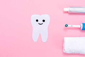 A silhouette of a tooth cut out of felt with a smiling cartoon face, next to an electric toothbrush, towel and toothpaste. Pink background. Hygiene of the oral cavity