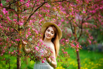 Beautiful young red-haired woman enjoys the fragrance in the spring blooming garden against the background of flowering trees. Young dreamy smiling lady in nature at sunset. 