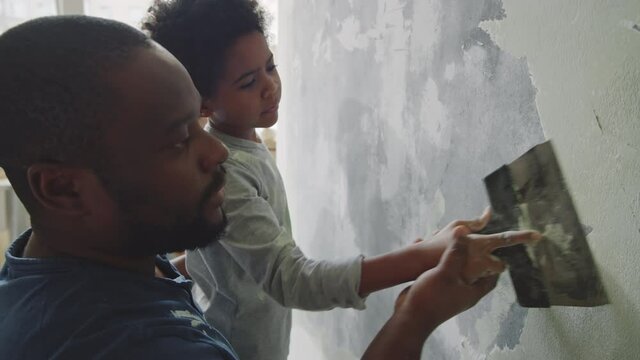 Afro-American father teaching little son how to scrape wall with paint scraper while renovating home together