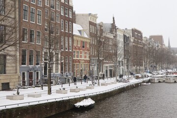 Snowy Amsterdam Canal View with Historic Buildings, Boats, Trees and People Walking