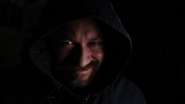 A man in a hood smiles viciously, unkindly on a black background.