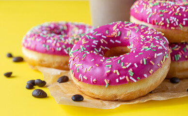 Yummy pink glaze donuts with colorful sprinkles, takeaway coffee cup and roasted coffee beans on yellow background. Junk food. Close up.