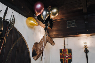 On a light gray wall next to a torch-shaped lantern, a medieval knight's shield and a sword hangs a stuffed antelope head and neck with balloons. Birthday and holiday hunting inn concept.