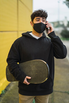 Chico joven atractivo con mascara por covid 19 con skate en una pared amarilla