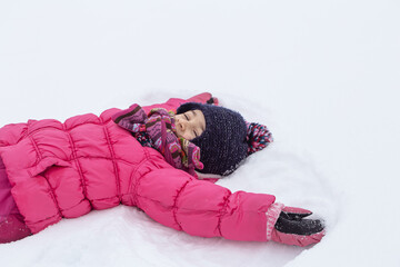 Little girl in a pink jacket and hat lies in the snow copy space.