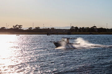 Small boat drives into sunset with spray reflecting light and shore and mountains in the distance