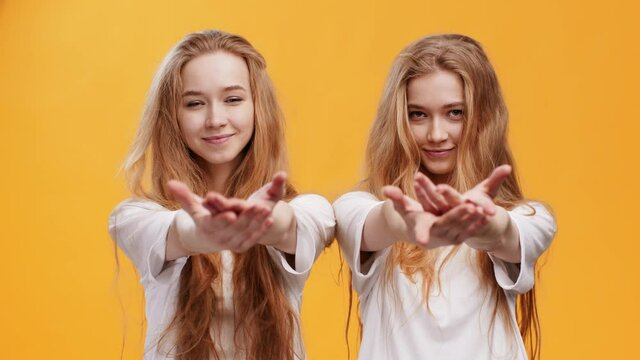 Gratitude concept. Two young redhead twin sisters touching chests and reaching hands to camera, orange background
