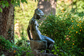 Nymph in woods - Garden statue of nude woman sitting on stump hidden among the trees and flowers in...