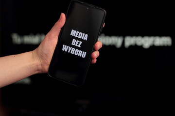 Media blackout protest in Poland. A person is holding a smartphone with the sign “Media bez wyboru” (Eng. Media without a choice) in front of a black TV screen. - obrazy, fototapety, plakaty