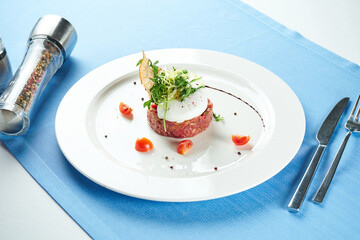 Delicious appetizer - beef tartare with croutons, poached egg and rucola salad in a white plate on a blue tablecloth