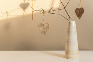 Creative composition in neutral colors for Valentine's Day. Hearts on a branch in a vase on the table. Handmade, coarse burlap fabric. Warm home atmosphere, romantic greeting card with space for text