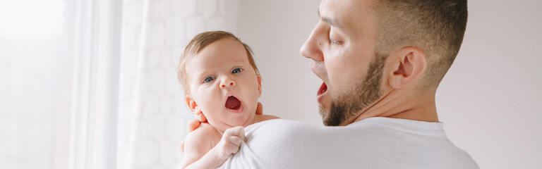 Fototapeta premium Fathers Day holiday. Caucasian father holds yawning newborn baby. Authentic lifestyle candid funny moments. Single young dad family life concept. Web banner header.