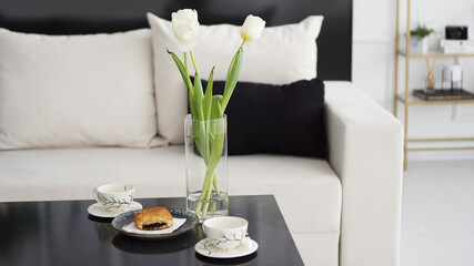 Sofa in a modern interior. On the table are a bouquet of tulips, coffee cups and a sweet bun. Black and white interior and sunny mood.