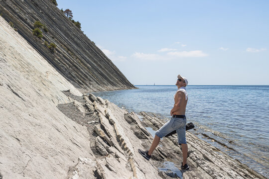 Summer sea landscape. The guy walking along the stone wild beach stopped and looks up. On the horizon, clear blue sky and steep cliffs. The resort city of Gelendzhik. Russia, Black Sea coast