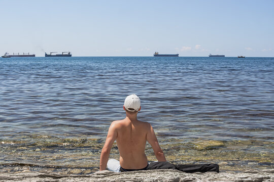 Summer sea landscape. A guy sits on a stone wild beach and looks into the distance. On the horizon is a clear blue sky and cargo ships. The resort city of Gelendzhik. Russia, Black Sea coast