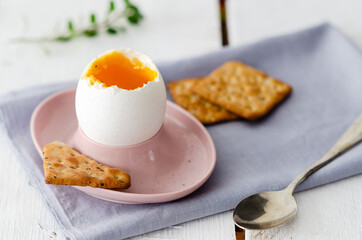 Soft boiled egg in a pink egg cup with a spoon, a light blue napkin, seed crackers, and oregano leaves, in white wooden backdrop. 