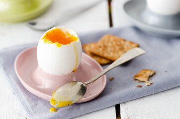 Soft boiled eggs in pink, light blue, and green egg cups with spoons, a light blue napkin, and seed crackers, in white wooden backdrop. 