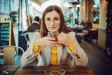 Young smiling fashionable brunette sitting on terrace of a bar, holding cup of tea and looking at camera.