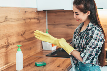 Girl puts on yellow rubber gloves before cleaning wooden kitchen countertop in the afternoon. Home. Cleaning. The girl in a plaid shirt and blue jeans. Girl washes the kitchen