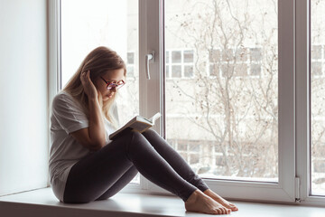 A young blonde beautiful woman in the white t-shirt and grey leggings with glasses is sitting on...