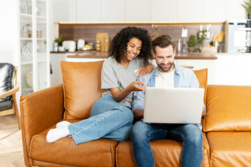 Cheerful diverse couple in love spends leisure time online with a laptop at home. Young woman and...