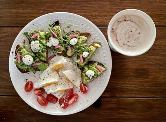 Overhead view of avocado toast with microgreens, fresh mozzarella, pickled turnips, eggs, tomatoes, and latte with wooden table background