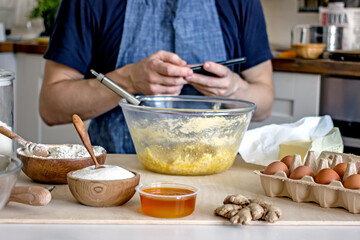 A faceless man in an apron kneads cookie dough with honey, flour and butter on the kitchen table with ingredients. Authentic hobby home baker. Close-up of a glass cup with dough and hands with a whisk