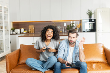 A multiracial couple with joysticks sits on the couch at home and playing video game, an African woman is winner, scream happily while her boyfriend feels upset with a losing