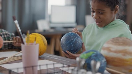 Panning medium close-up of cute afro siblings chatting while crafting and painting diy globe models sitting together at table in living room